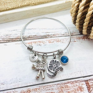 Può includere: Bracciale a cerchio color argento con ciondoli a tema cinematografico. I ciondoli includono una bobina di film, una cinepresa vintage, un cristallo blu e un disco con la lettera "H". Il bracciale è esposto su una superficie di legno bianca e marrone.