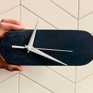 Pode incluir: Um relógio moderno e minimalista com um corpo de madeira ovalado azul marinho escuro. O relógio apresenta ponteiros brancos e um mostrador branco. Os ponteiros apontam para as 10h10. O relógio é segurado em frente a um fundo de azulejos brancos.