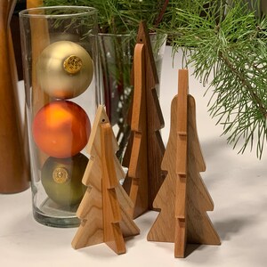 May include: Three wooden Christmas tree ornaments, each with a different design. The trees are made of light brown wood and have a natural finish. The trees are standing on a white surface.