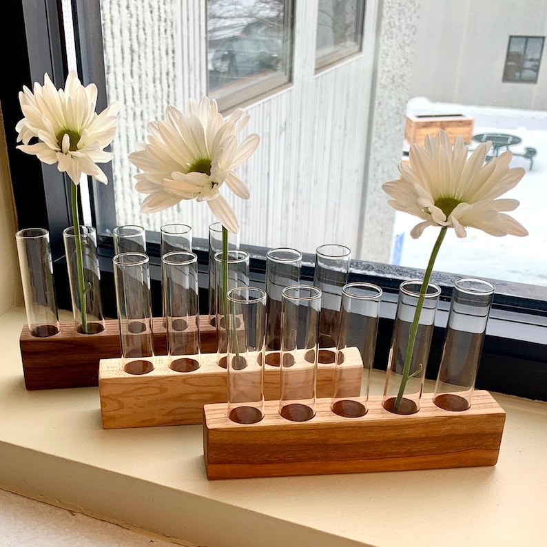 May include: Three wooden flower vases with glass test tubes for holding flowers. The vases are made of light brown wood and have a natural finish. The test tubes are clear glass and are arranged in rows of three. The vases are filled with white daisies.