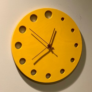 Könnte beinhalten: Eine gelbe runde Uhr mit goldenen Zeigern und einem Kreisdesign um das Zifferblatt.