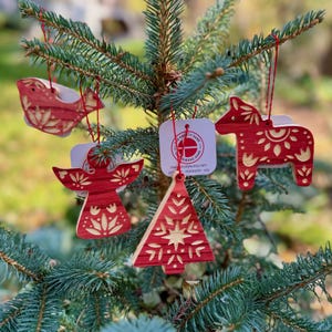 May include: Red wooden Christmas decorations in the shapes of a bird, angel, horse, and Christmas tree, hanging on a fir tree. Each ornament features detailed cut-out patterns. The decorations are suspended by red strings.