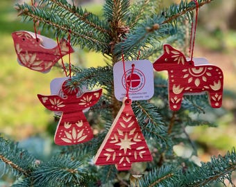 Puede incluir: Adornos navideños de madera roja con forma de pájaro, ángel, caballo y árbol de Navidad, colgados de un abeto. Cada adorno tiene intrincados diseños recortados. Los adornos están suspendidos por cuerdas rojas.