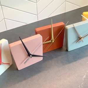 May include: Five square-shaped clocks with different colored faces. The clocks are arranged in a row from left to right. The colors of the clocks are white, pink, orange, light blue, and yellow.