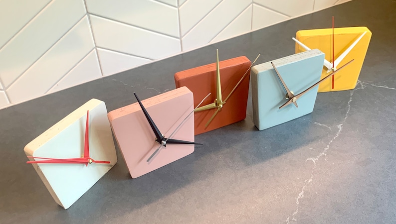 May include: Five square-shaped clocks with different colored faces. The clocks are arranged in a row from left to right. The colors of the clocks are white, pink, orange, light blue, and yellow.