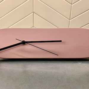 May include: A pink oval clock with black hands and a gold center. The clock is sitting on a gray surface.