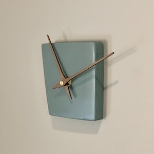 May include: A square, light blue clock with wooden hands. The clock is mounted on a white wall.