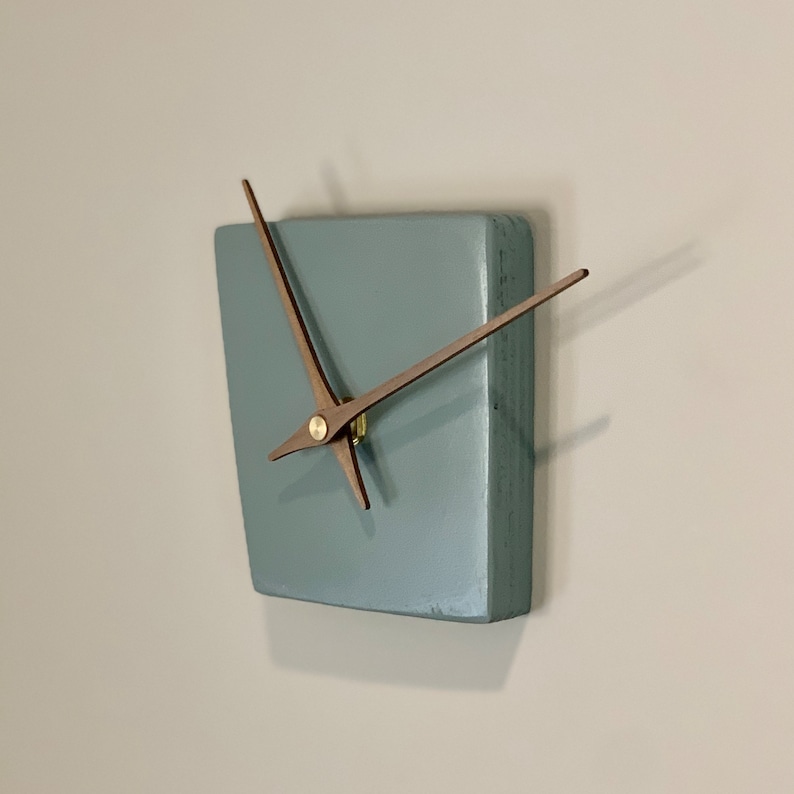 May include: A square, light blue clock with wooden hands. The clock is mounted on a white wall.