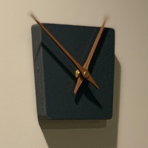 May include: A square, dark gray clock with wooden hands. The clock is mounted on a wall.