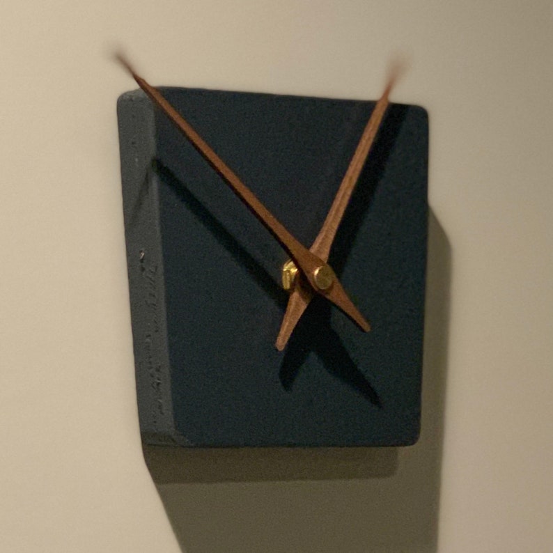 May include: A square, dark gray clock with wooden hands. The clock is mounted on a wall.
