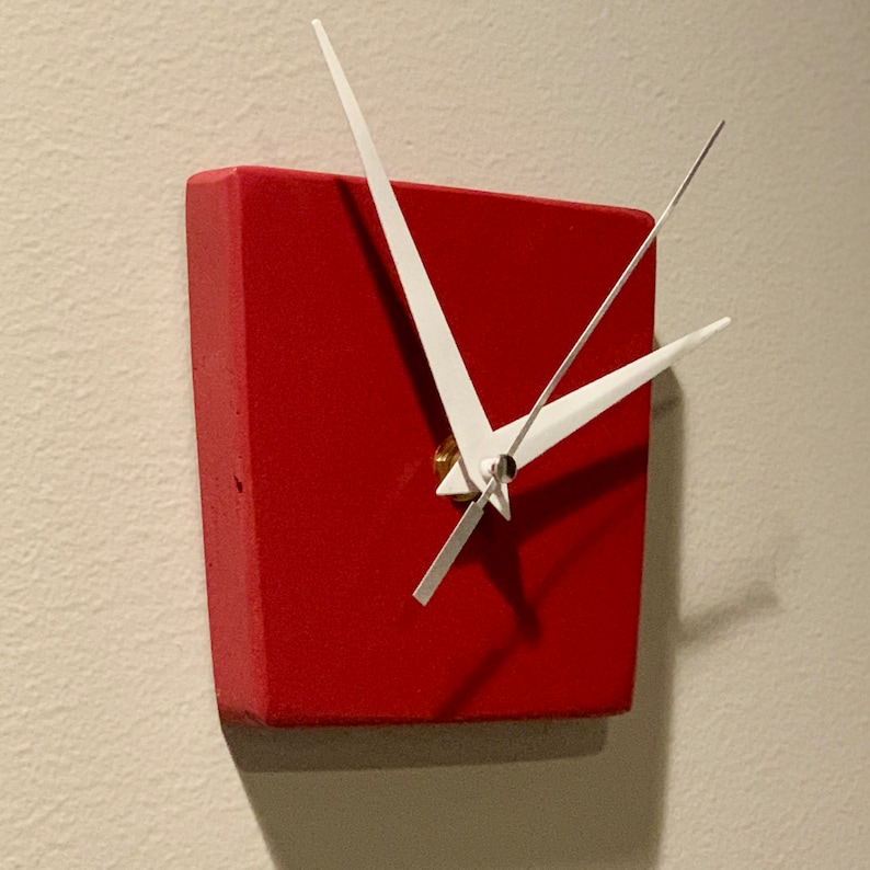 May include: A red square clock with white hands. The clock is mounted on a white wall.