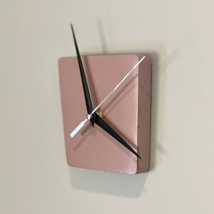 May include: A square, blush pink clock with black hands and a gold center. The clock is mounted on a white wall.