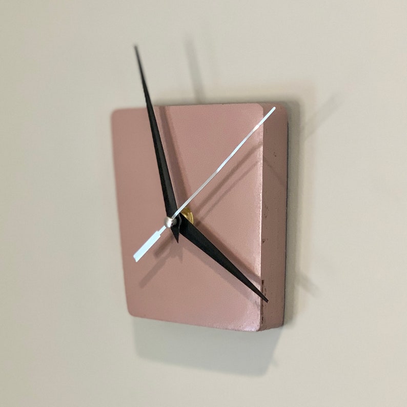 May include: A square, blush pink clock with black hands and a gold center. The clock is mounted on a white wall.