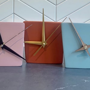 May include: Five square clocks with different colored faces and hands. The clocks are white, pink, orange, light blue, and yellow.