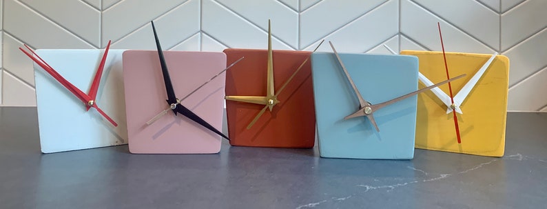 May include: Five square clocks with different colored faces and hands. The clocks are white, pink, orange, light blue, and yellow.