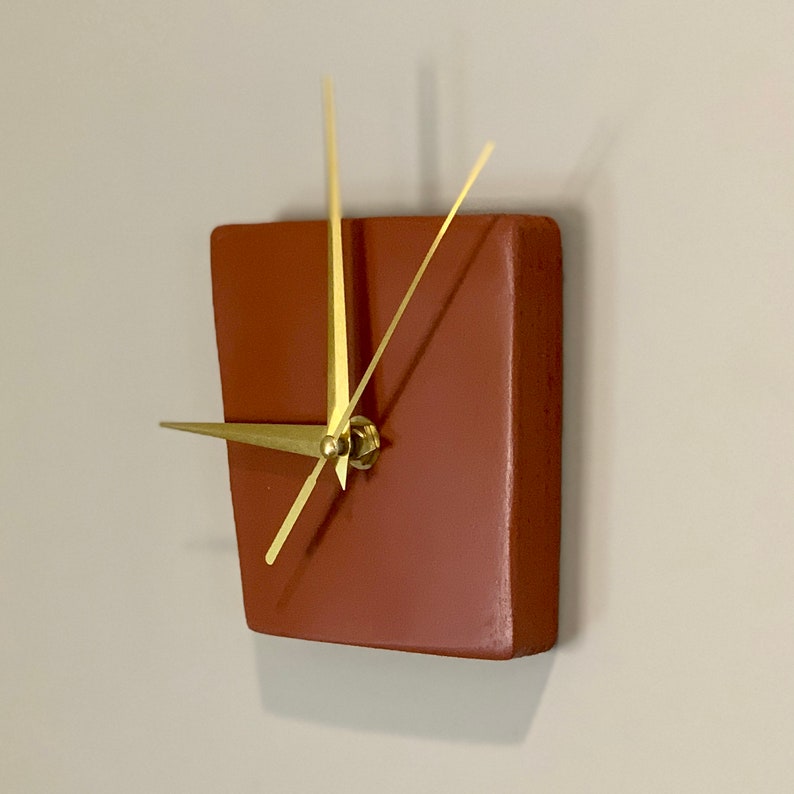 May include: A square terracotta-colored clock with gold hands. The clock is mounted on a wall.