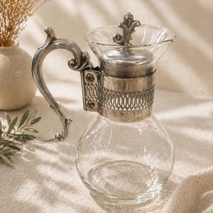 May include: A clear glass carafe with a silver metal handle and accents. The carafe has a round base and a flared top with a decorative lid. The handle is ornate, and the metal band around the carafe is detailed. The carafe is likely for serving beverages.