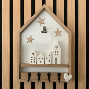 Puede incluir: Un organizador de pared de madera con forma de casa que incluye un panel trasero blanco, cuatro figuras de casas blancas sobre un estante, tres estrellas de madera, un clip metálico, cuatro ganchos negros y un pequeño corazón blanco.