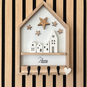 Può includere: Portachiavi in legno a forma di casa, con un ripiano con case bianche in miniatura e una fila di ganci neri. Il design include stelle di legno e il testo "Zuhause voller Liebe".