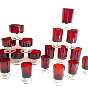 Op de afbeelding: Een verzameling vintage robijnrode glaswerk, waaronder dessertschalen met voet en tumblers. De glazen zijn gerangschikt in een piramide en verspreid over een witte achtergrond. Het glaswerk is gemaakt van dieprood glas met heldere stelen.