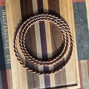 May include: A thick, copper rope, braided into a circular shape, is displayed on a wooden cutting board with a striped pattern. The board is on top of a fabric with a red, white, and blue pattern.