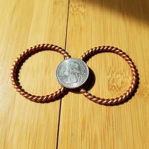 May include: Two copper wire rings, twisted and intertwined, with a US quarter dollar coin resting in the center. The coin is positioned so that the words "United States of America" and "Liberty" are visible.
