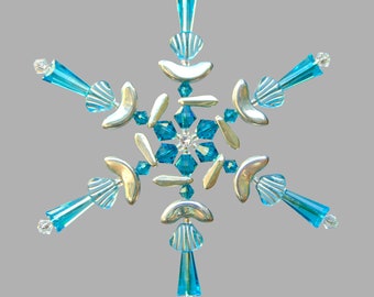 May include: A handcrafted snowflake ornament with a central cluster of blue crystals and a silver-colored gem. The six points feature turquoise-colored glass beads and silver-colored crescent-shaped accents. The ornament is suspended by a small loop.