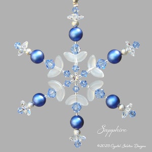 May include: A snowflake ornament made of blue and clear beads and white glass. The ornament is hanging from a silver string and has the word "Sapphire" written below it.