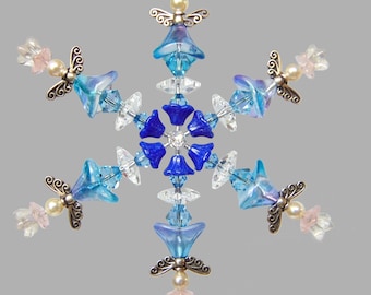 May include: A snowflake ornament made of blue, white, and pink glass beads. The snowflake has six points and is decorated with small angels. The ornament is hanging from a silver string.