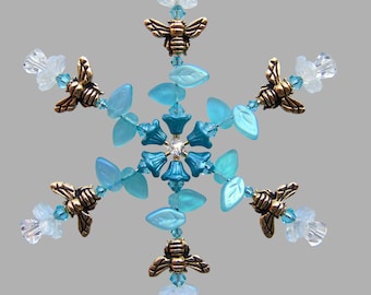 May include: A snowflake ornament with a central flower design, surrounded by bronze-colored bee charms and light blue leaf-shaped beads. Clear and light blue crystal beads accent the design. The ornament is suspended by a gold-colored wire.