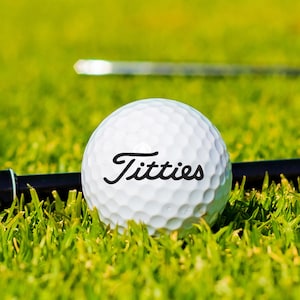 May include: A white golf ball with the word "Titties" printed on it in black cursive lettering. The ball is sitting on green grass.