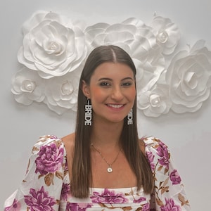 May include: A woman with long brown hair is wearing a white and purple floral blouse. She is wearing a gold necklace with a small round pendant. She is also wearing long, silver earrings that spell out "BRIDE".