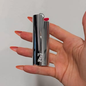 May include: A silver-colored lighter with a red ignition button. The lighter has a decorative design of the letter "Z" embellished with rhinestones. The lighter is held in a hand with long, orange-colored nails.