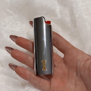 May include: A silver-colored lighter with a red ignition button. The lighter has a gold-colored emblem on the front. The lighter is held in a hand with long, polished nails.