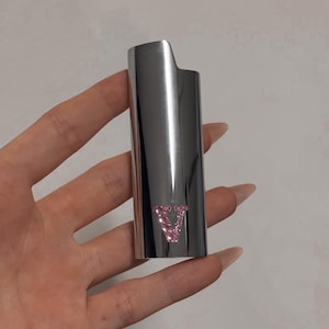 May include: A silver-colored lighter with a pink, jewel-encrusted "V" logo. The lighter has a sleek, metallic finish and a rectangular shape with a curved top. The pink logo adds a touch of sparkle to the design.
