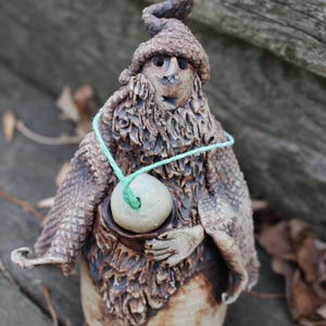 May include: A ceramic figurine of a bearded gnome wearing a hat with a long, curved point. The gnome is holding a round, brown object in its hands. A green string is tied around the gnome's neck and the object.