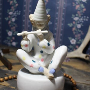 May include: A ceramic figurine of a seated clown playing a flute. The clown is dressed in a white outfit with colorful polka dots and a tall, conical hat. The figurine sits on a white base.
