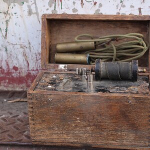 May include: A vintage wooden box containing a metal spool and two brass cylinders. The box is open and the spool is visible. The box is made of wood and has a worn finish.