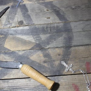 May include: A curved metal sickle with a wooden handle lies on a weathered wooden surface. A silver cross necklace and a handprint are also visible. The background features a faded, dark design.