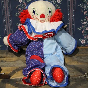 May include: A vintage clown doll with a cream-coloured face, red yarn hair, and a blue and white ruffled collar. The doll's outfit is half blue with a floral pattern and half solid light blue, with red polka-dot feet.