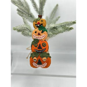May include: A stacked pumpkin ornament with three orange pumpkins, each with a carved face. The top pumpkin is yellow. Green leaves and a brown string are attached. The ornament is hanging in front of a green, leafy background.
