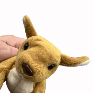 Peut inclure: Un jouet en peluche kangourou brun et blanc avec une étiquette en forme de cœur rouge. Le kangourou est assis, la tête inclinée sur le côté et les yeux ouverts.