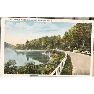 May include: A vintage postcard depicting a scenic view of Deerfield River along the Mohawk Trail. The postcard shows a winding road with a white wooden fence along the edge of the river. The river is surrounded by lush green trees and a blue sky with white clouds.