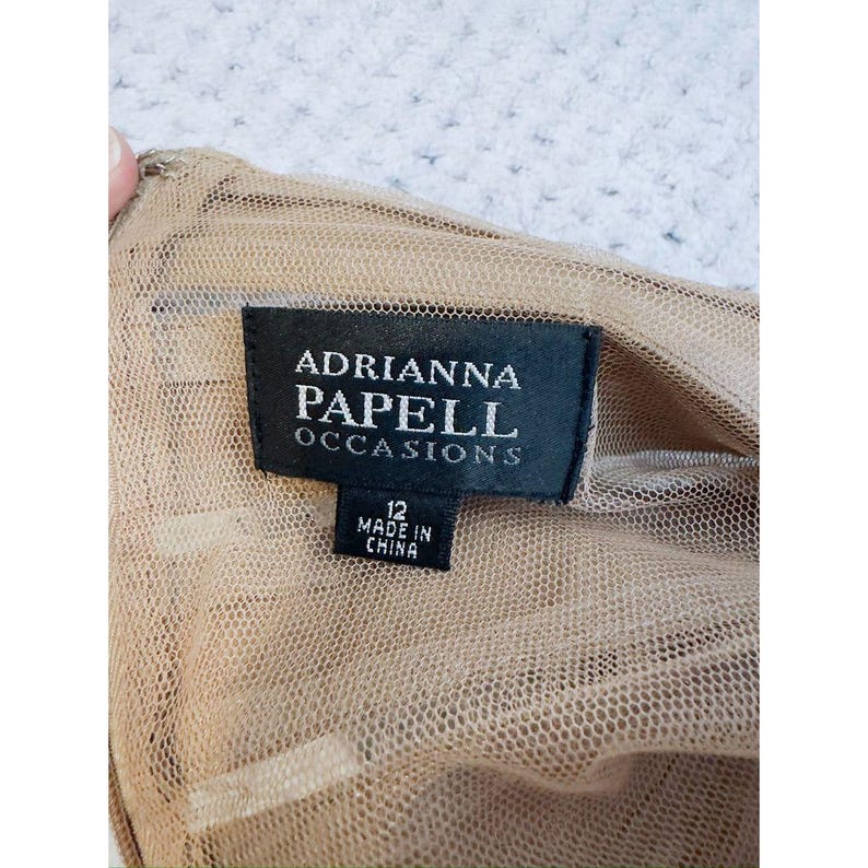 Może przedstawiać: Zbliżenie metki od Adrianna Papell Occasions. Czarna metka z białym napisem "ADRIANNA PAPELL OCCASIONS" i "12 MADE IN CHINA". Metka jest przymocowana do beżowej siateczkowej tkaniny.