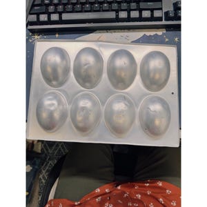 May include: A rectangular, aluminum baking mold with eight oval-shaped cavities. The mold is designed for creating egg-shaped treats or desserts. The metal has a slightly worn appearance, suggesting previous use. The mold sits on a surface with a keyboard in the background.