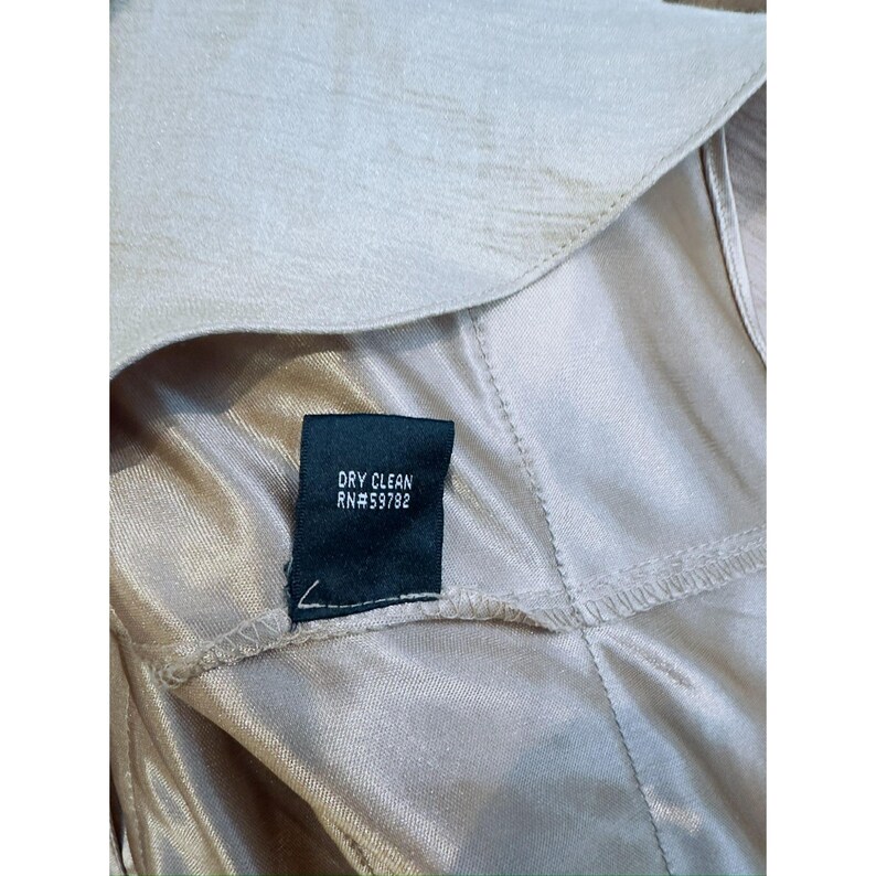 Może przedstawiać: Zbliżenie beżowego ubrania o satynowej fakturze. Ciemnozielona metka z białym napisem "DRY CLEAN RN#59782". Ubranie wygląda na sukienkę lub top, z widocznymi szwami i kołnierzem.