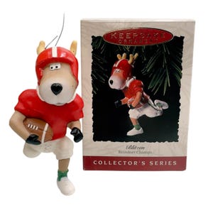 May include: A Christmas ornament featuring a reindeer in a red football uniform and helmet, holding a football. The ornament is next to a box with a matching illustration and the text "Blitzen Reindeer Champs".