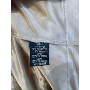 Może przedstawiać: Zbliżenie metki z informacjami o składzie materiału. Na metce widnieje napis "SHELL 71% RAYON 18% POLYESTER 11% NYLON COMBO 100% POLYESTER LINING 100% POLYESTER MADE IN CHINA". Etykieta jest czarna z białym tekstem.