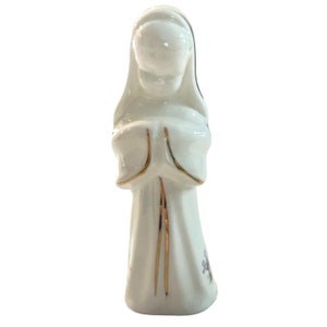 May include: A white porcelain figurine of a woman in a long white robe with gold trim and purple flowers. She is holding her hands together in prayer.