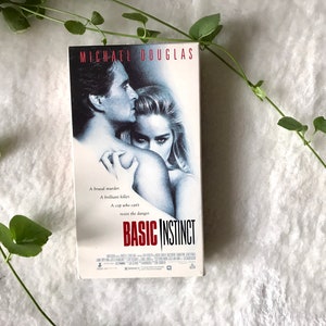 May include: A VHS tape for the movie "Basic Instinct" featuring Michael Douglas and Sharon Stone. The cover art is a black and white image of the actors. The title is in red and white. Green vines and leaves are in the background.